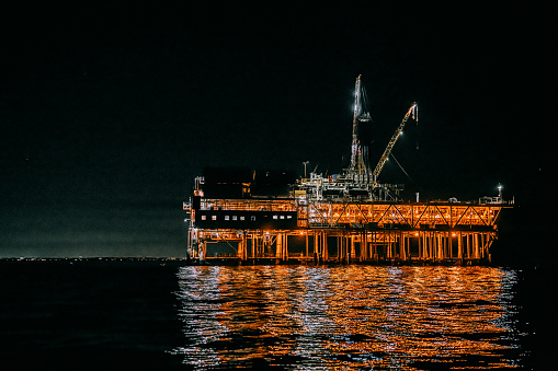 An epic, cinematic, dark photograph of offshore oil drilling at night off the coast of Huntington Beach, California. The industrial machinery and equipment used in the drilling and extraction of fossil fuels are highlighted against the night sky. 
This image speaks to issues of fuel and power generation and environmental concerns surrounding the oil and gas industry.