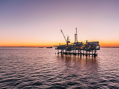 A stunning image of an offshore oil rig at sunset off the coast of Huntington Beach, California. The setting sun highlights the industrial machinery and equipment used in the drilling and extraction of fossil fuels, including crude oil and natural gas. 

This photograph captures the intersection of the energy industry and the natural beauty of the Pacific Ocean, and speaks to issues of fuel and power generation, energy crises, and environmental concerns surrounding the oil and gas industry.