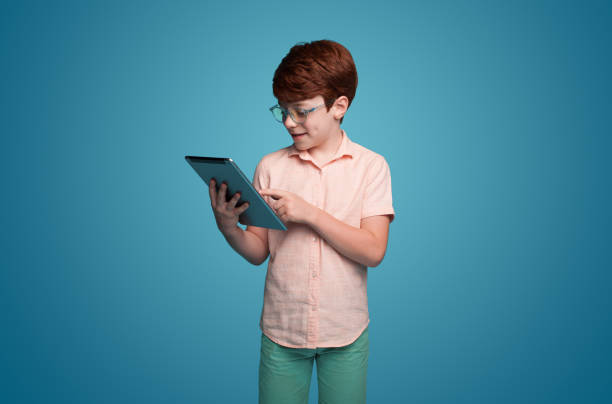 Cute schoolboy wearing eyeglasses holding digital tablet for online learning isolated over blue background. Education internet technology. Modern lifestyle. Online education. stock photo