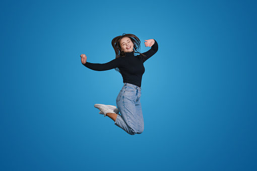 Portrait of cheerful positive brunette woman jumping in the air with raised hands isolated on blue background. Life people energy concept