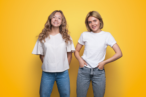 Portrait of two cheerful young women standing together looking at camera isolated over yellow background.