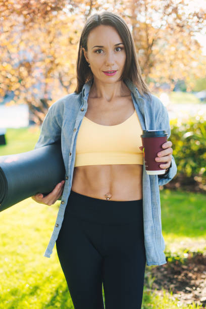 Attractive athletic woman instructor in sportswear standing in green summer park with rolled mat and plastic cup of coffee, having rest after training lesson. Take a break. Workout sport concept. Fitness woman. Health care concept. stock photo