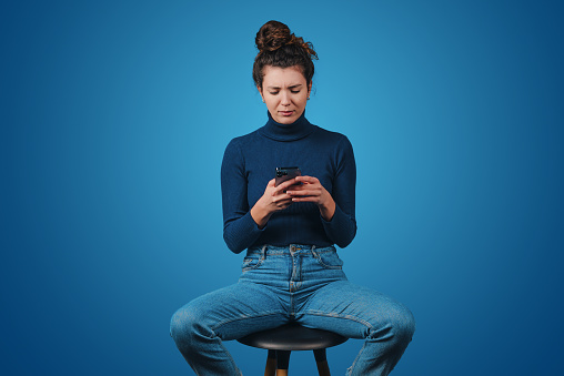 Photo of young pretty woman sitting on the bar stool and holding a smartphone isolated over blue background. Millennial lifestyle. Social media.