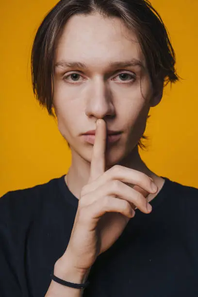 Handsome man putting his index finger to his lips, tries to keep conspiring. Shh, be quiet please. Isolated shot of attractive man on yellow background.