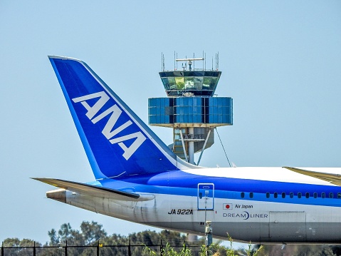 An All Nippon Airways Boeing 787-9 taxiing past the air traffic control tower at Sydney Kingsford-Smith Airport and heading to the southern end of the main north-south runway.  The plane, JA922A, flight number NH890, was flying to Tokyo.  This image was taken from Kyeemagh Avenue, Kyeemagh on a sunny afternoon on 26 February 2023.