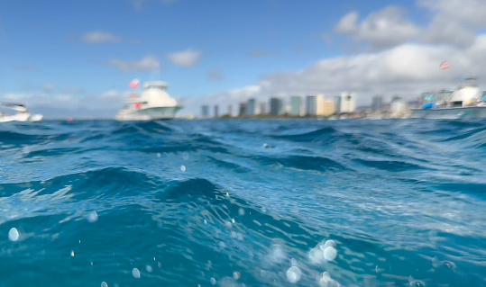 Ripples, waves, and water dropplets over the Pacific ocean with boats and buildings on shore out of focus in the background. Honolulu, Hawai'i.