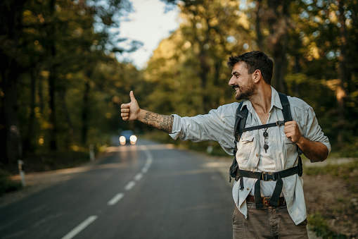 A man walking through a forest wearing a casual outfit and a backpack and hitchhiking. Happy man on a holiday hiking in a countryside location.