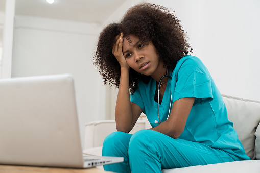 Stressed female doctor with hand on forehead while working with laptop computer in hospital. Unhappy female nurse in office at hospital