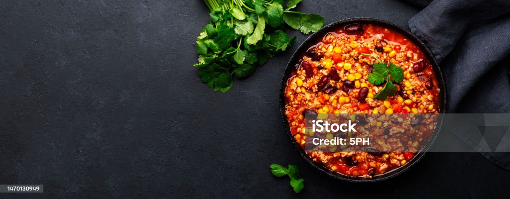 Chili con carne with beef, red beans, paprika, corn and hot peppers in tomato sauce, spicy tex-mex dish in a cooking pot, black table background, top view banner Chili Con Carne Stock Photo