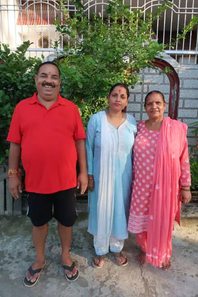 Photo of Image of Hindu, Indian two generation family, husband, wife and daughter, man in casual clothing, women dressed in Salwar Kameez traditional clothing, wearing sindoor in hair parting, bindi on forehead, full length view, standing in paved yard