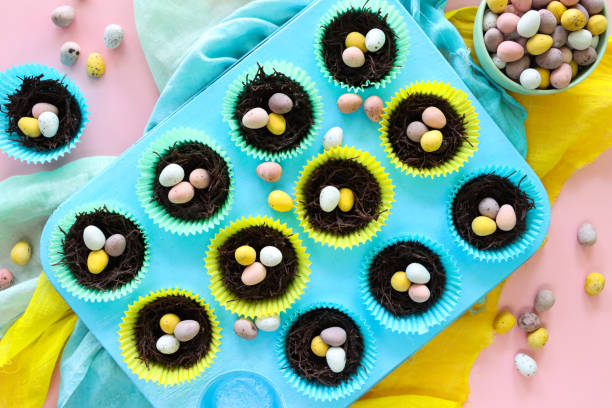 Image of muffin baking tray with rows of homemade chocolate bird nest cakes in brightly coloured paper cake cases, topped with mini chocolate Easter eggs, fluffy yellow chick toys decoration, yellow, green and turquoise muslin, pink background Stock photo showing close-up, elevated view of baking tray lined with multi-coloured paper cake cases containing Easter cake made of chocolate and cereal bran threads in the form of bird nests decorated with mini chocolate eggs. muffin tin eggs stock pictures, royalty-free photos & images