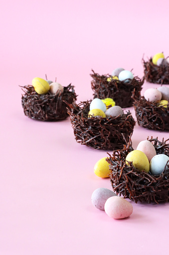 Stock photo showing close-up view of pink surface covered with Easter cakes made of chocolate and cereal flakes in the form of bird nests decorated with mini chocolate eggs and fluffy yellow chick toys.