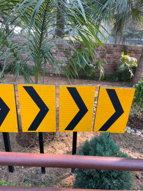 Close-up image of chevron road signs showing a curve , palm trees, red brick wall background, focus on foreground Stock photo showing close-up view of row of yellow and black chevron road signs advising a curve to keep the road safe. road chevron warning signs stock pictures, royalty-free photos & images