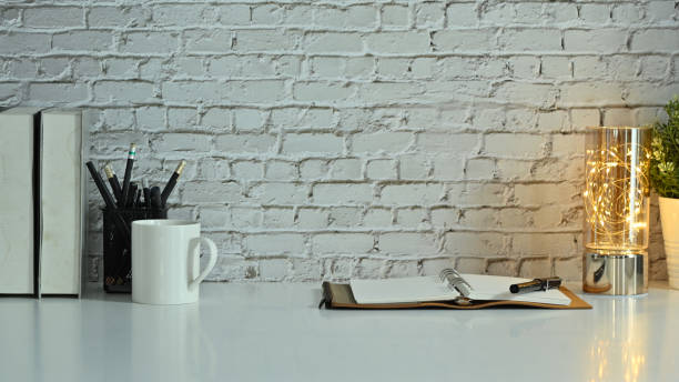 istock Stylish workplace with opened notebook, pencil holder, coffee cup and books on white table against brick wall. 1470144278