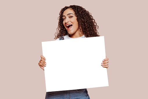 Young cheerful mixed race woman with curly hair, holding blank banner at studio, isolated over beige background. Advertising, announcement, offer.