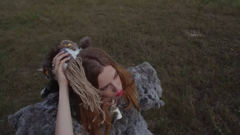 Close up portrait  of blonde girl in a pagan animal-like  fur  made of skins standing  against the grass backdrop taking of a goat mask with horns and looking up at the sky