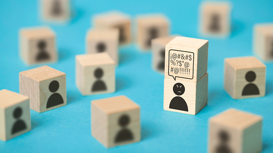 Front view of a group of  wooden cubes with icons representing persons. The main focus is on an angry person who has a cube over his head with a thought bubble full of insults