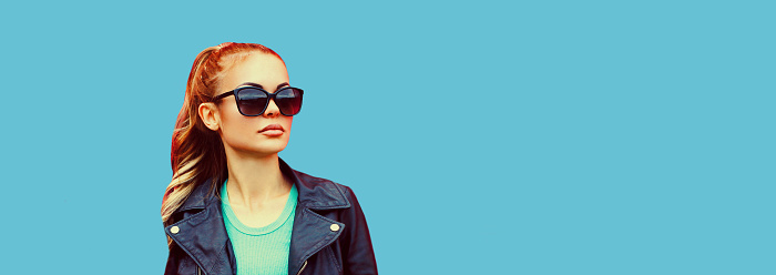 Portrait of beautiful blonde young woman wearing sunglasses, black rock jacket on blue background, blank copy space for advertising text