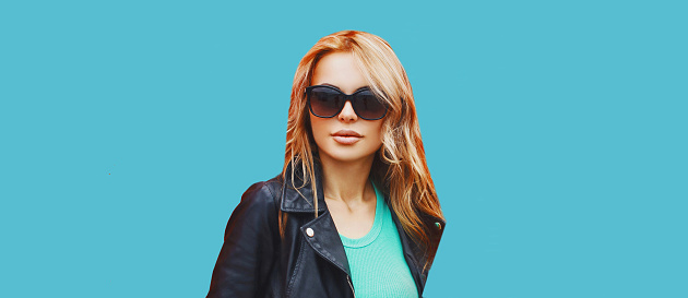 Portrait of beautiful blonde young woman wearing sunglasses, black rock jacket on blue background