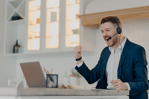 Business success and achievement. Excited happy businessman in formal wear suit and headset celebrating success, raising clenced fist and looking at laptop while sitting at his workplace at home