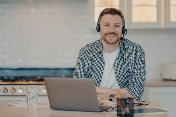 istock Handsome glad man customer support manager gives advice or consultation to clients works online from home poses at home kitchen wears wireless headphones uses modern laptop computer. Remote job 1470204174