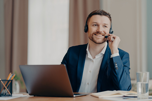 Photo of happy male manager wears headset works on laptop computer consults clients looks with glad thoughtful expression away dressed formally poses at desktop. Support and online consultation