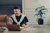 Happy bearded young man studies indoor with modern gadgets writes down notes from internet website makes records in notebook holds smartphone poses at coworking space cozy interior. Hipster student