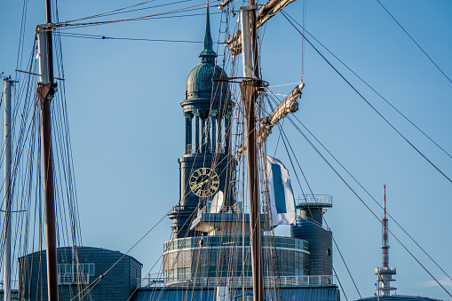 The beautiful clock tower of St. Michaelis Church and TV-tower Heinrich-Hertz-Turm are captured through the rigging of a sailboat, showcasing the maritime heritage of Hamburg in the evening sun.