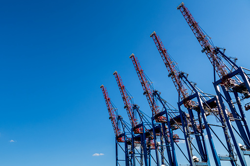 Towering red and blue cranes against blue sky, emphasizing the role of harbor facilities and infrastructure in the world economy and global freight transportation, with copy space on the left side.