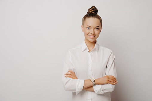 Beautiful female company worker with charming smile in formal white shirt posing in studio with crossed arms, standing isolated on grey background. Businesswoman being in good mood at work
