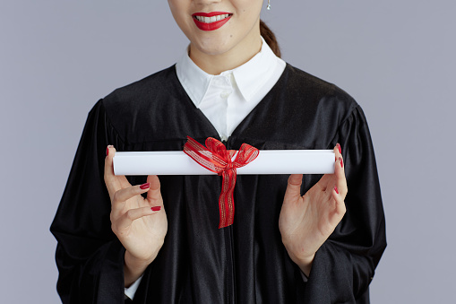 Headshot of a teenage girl at her high school graduation wearing cap and gown, standing outdoors with her mother, smiling at the camera. The graduate is Hispanic and White.