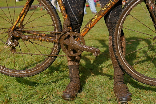 Close up of orange and yellow bicycle covered in mud with rider's legs covered in mud behind the bike frame. Crank set is in the middle of frame. Green grass in background.