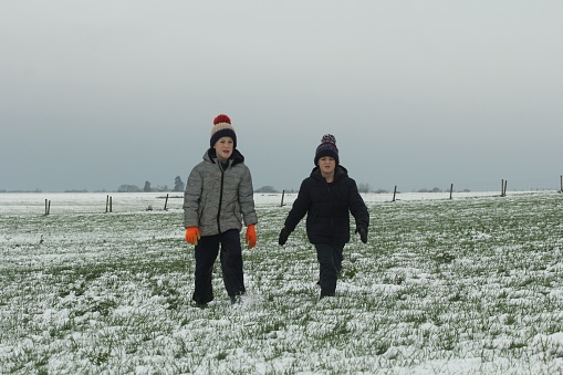 Two young boys walking through grass field with light covering of snow. They are wearing warm winter clothing, the sky is grey and overcast