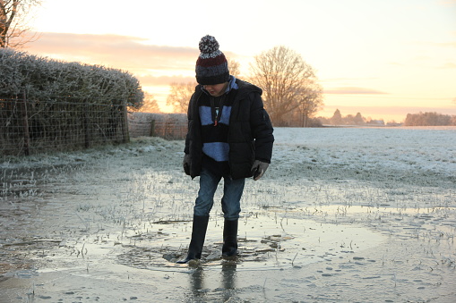 Young boy wearing warm winter clothing stepping on and breaking through icy puddle in snowy field