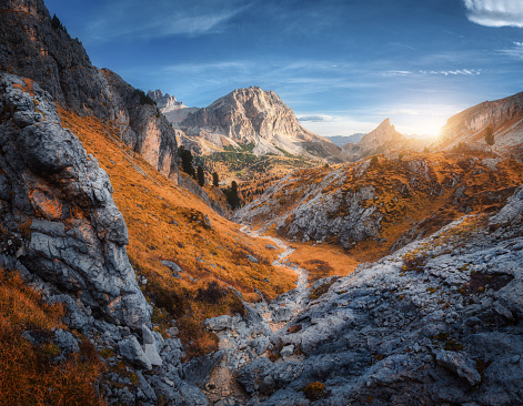 Beautiful mountain path, rocks and stones, orange trees at sunset in autumn in Dolomites, Italy. Colorful landscape with forest, rocks, trail, orange grass and blue sky in fall. Hiking in mountains