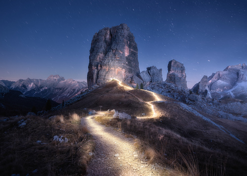 Flashlight trails on mountain path against high rocks at night in spring. Dolomites, Italy. Colorful landscape with light trails, trail on the hill, mountain peaks, purple sky with stars in summer