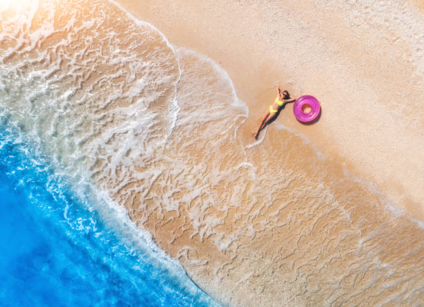 Aerial view of the lying beautiful young woman with pink swim ring on the sandy beach near sea with waves at sunny day. Summer in Lefkada island, Greece. Drone view of slim girl, clear blue water stock photo
