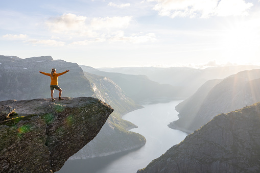 A woman stands while throwing her arms in the air on the mountain's cliff edge of Trolltunga throning over Ringedalsvatnet watching the sunset in the Norwegian mountains near Odda, Rogaland, Norway