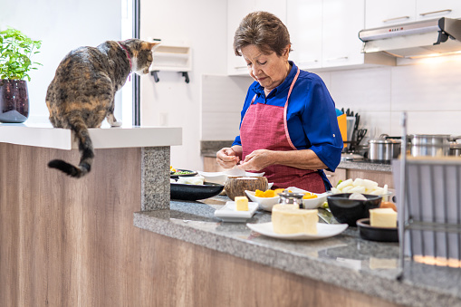 Elderly woman cooking in the kitchen while a tabby cat looks at her at the counter. Domestic life concept