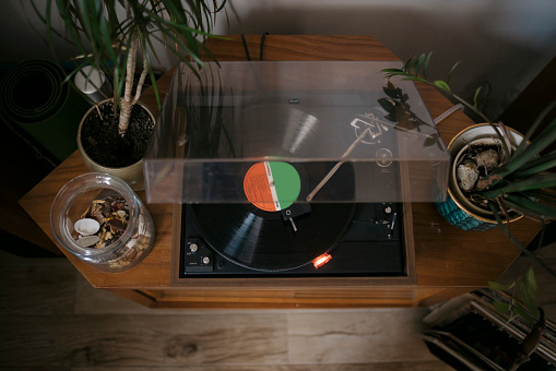 High angle view of a LP record player playing music at home. Old turntable device with LP disc playing retro music on side cabinet with potted plants in living room.