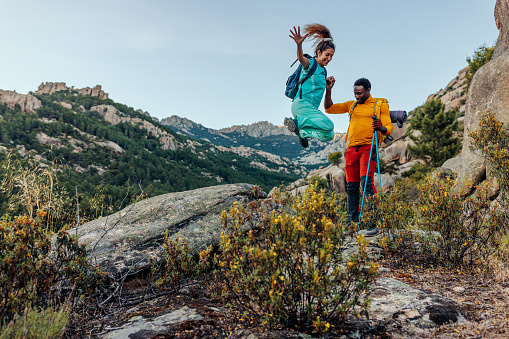 A young multiethnic couple is having fun during hiking. The man holds his girlfriends hand as she jumps of a rock filled with joy because of their time together.