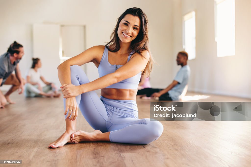 Woman sitting in a fitness studio with her yoga class Fit young woman smiling at the camera while sitting in a yoga studio with her class in the background. Happy young woman having a workout session with her class in a fitness studio. Women Stock Photo