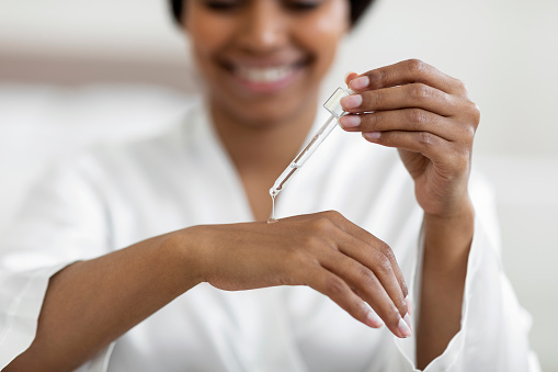 Smiling Black Woman Applying Moisturusing Serum On Hand With Dropper, Closeup Shot Of Attractive African American Female Testing New Beauty Product While Making Selfcare Treatments At Home