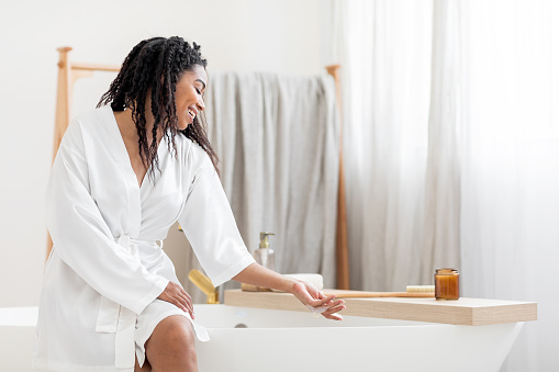 Home Spa. Beautiful Black Woman Sitting On Bathtub And Touching Foam, Attractive Young African American Female Wearing White Silk Robe Preparing To Take Bath, Relaxing In Bathroom At Home, Copy Space