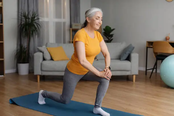 Positive sporty senior woman keeping fit at home, doing lunges, exercising on yoga mat in living room and smiling. Staying healthy at retirement