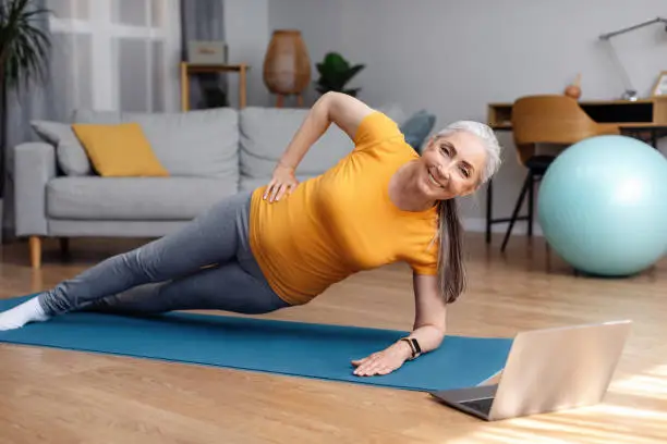 Photo of Happy fit senior woman standing in side plank, training on yoga mat in living room and using laptop
