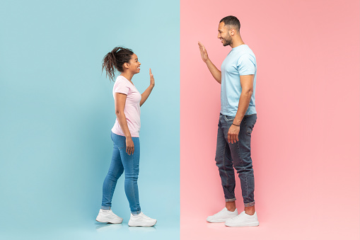 Side view of young black man and woman waving to each other or giving high five standing isolated on halved pink and blue studio background, greeting each other