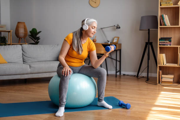 positive senior woman exercising with dumbbells, sitting on fitball and listening to music in headphones indoors - log fire audio imagens e fotografias de stock