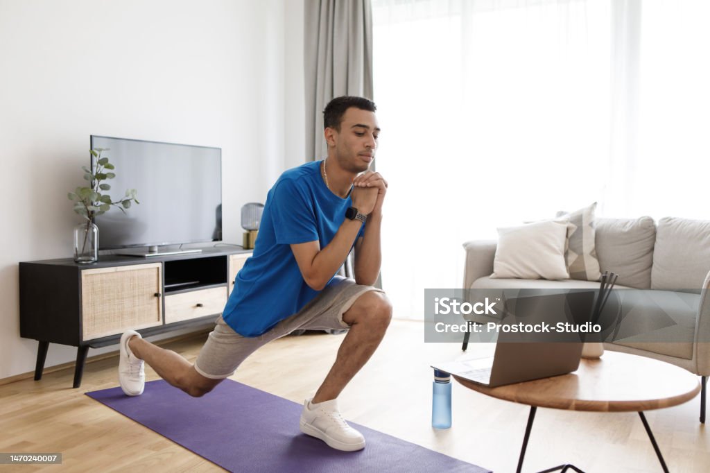Young arab man watching training tutorial on laptop and doing forward walking lunges on fitness mat, exercising at home Young arab man watching training tutorial on laptop and doing forward walking lunges on fitness mat, exercising at home. Focused guy looking at pc screen on tea table 20-24 Years Stock Photo