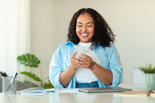 Happy Black Woman Using Mobile Phone Texting Sitting At Desk In Modern Office. Cheerful Female Using Application Via Cellphone At Workplace Indoor. Communication And Technology. Selective Focus